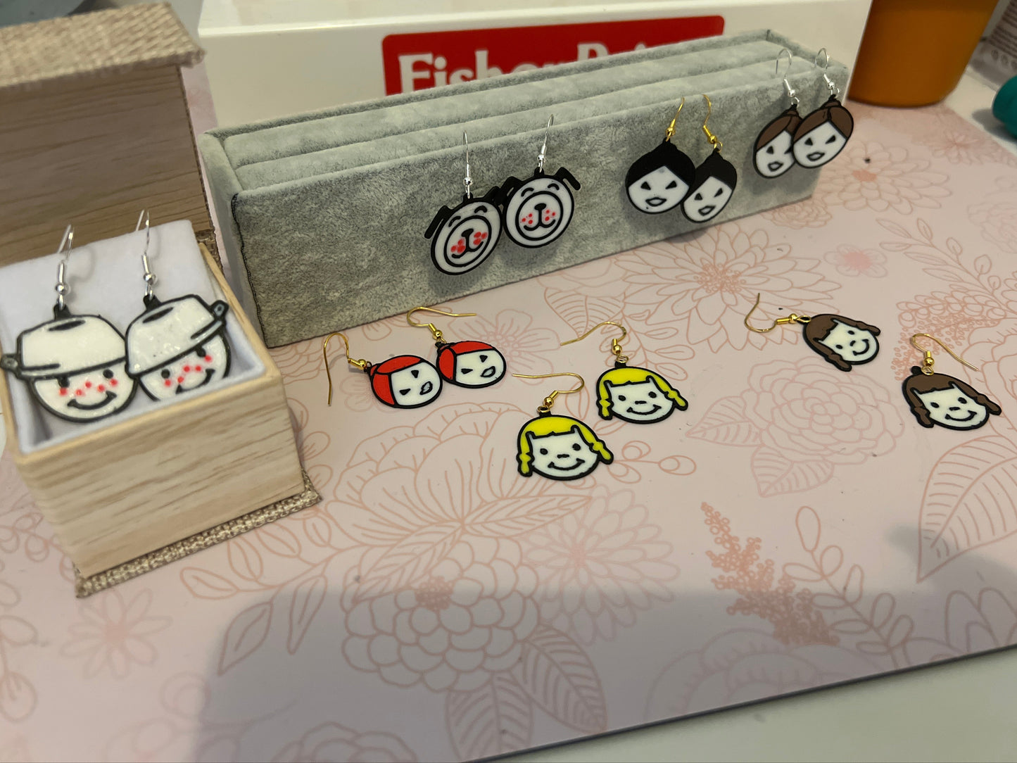 Set of cartoon character earrings displayed on a floral-patterned surface with a Fisher-Price box in the background.