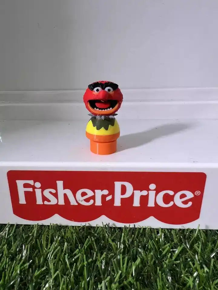 A custom Fisher-Price Little People figure of Animal from Sesame Street stands on a white surface over green grass.