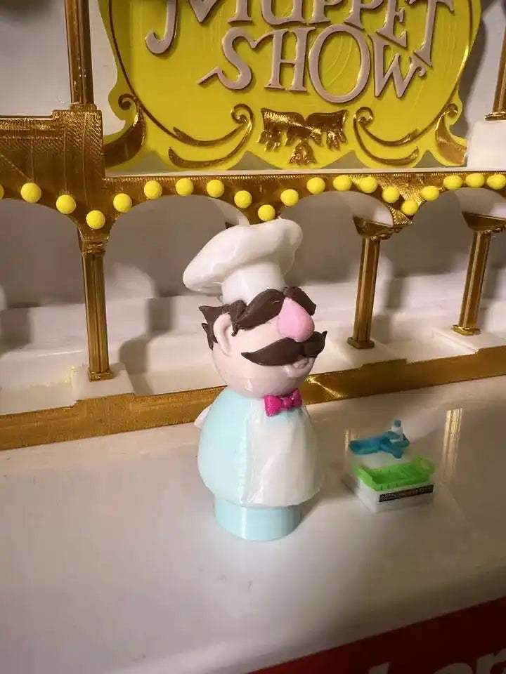 Chef puppet figurine on light blue base, white hat, pink bow tie, brown mustache