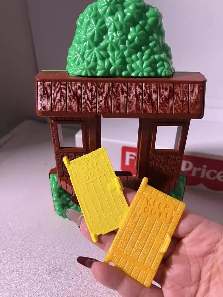 A hand holds two yellow replacement doors for the Fisher-Price #2551 Neighborhood Tree House, with "KEEP OUT!" on them.