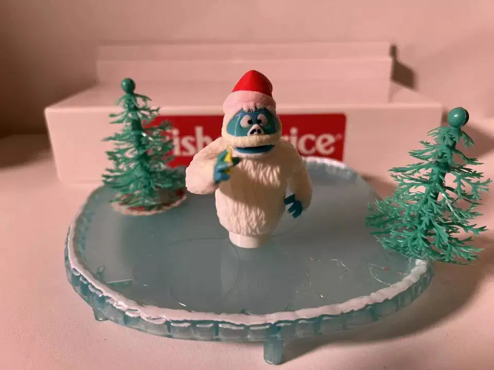 A custom 3D printed Little People-style "Christmas/Holiday Misfits - Yeti" figure, wearing a red Santa hat and holding a yellow item, stands on a translucent blue ice rink with two small green Christmas trees.