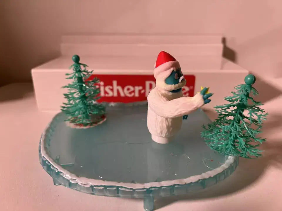 Holiday yeti figure in white fur with red Santa hat, waving beside green pine trees