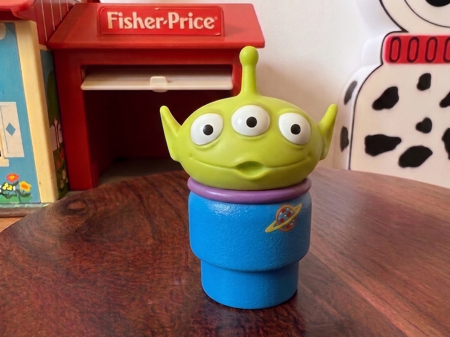Toy figure resembling a green alien with a blue body on a wooden surface, with Fisher-Price and Dalmatian-themed items in the background.