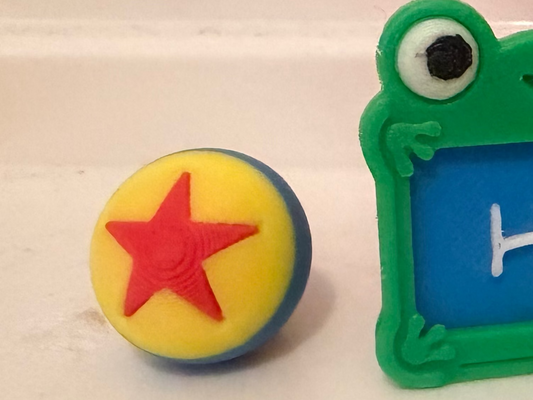 A colorful toy-like frog sign with a blue screen reading 'Hi!' and a green border, shown beside a yellow ball with a red star on a white surface. A large red and blue sign with yellow letters is blurred in the background.