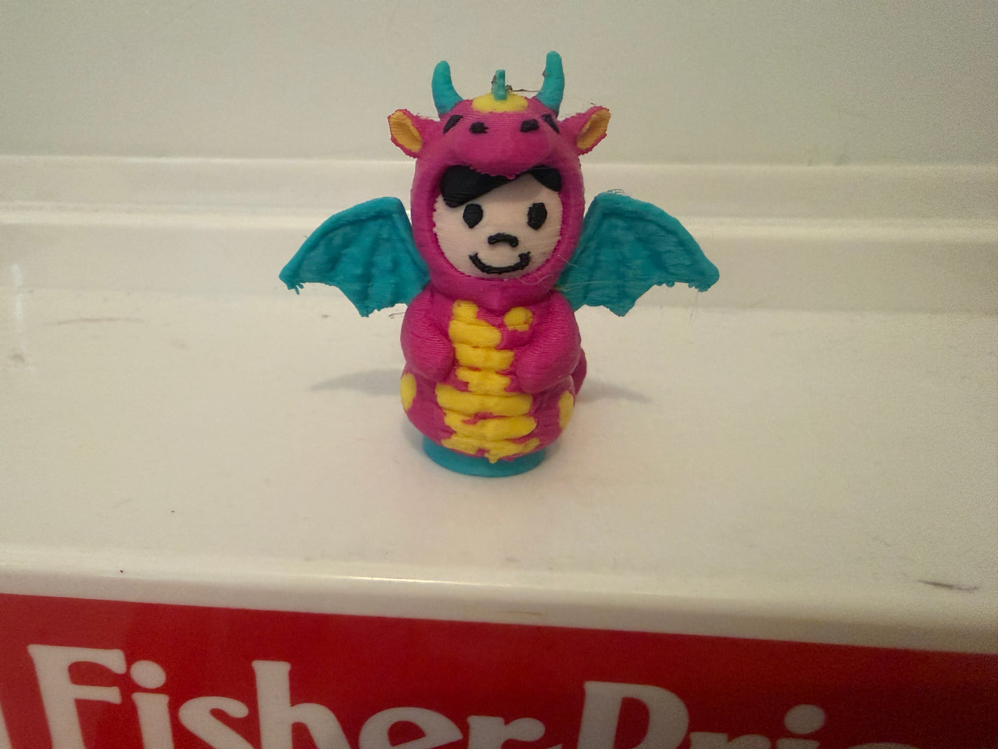 Pink dragon costume plush toy with teal wings and yellow belly, front-facing smile.
