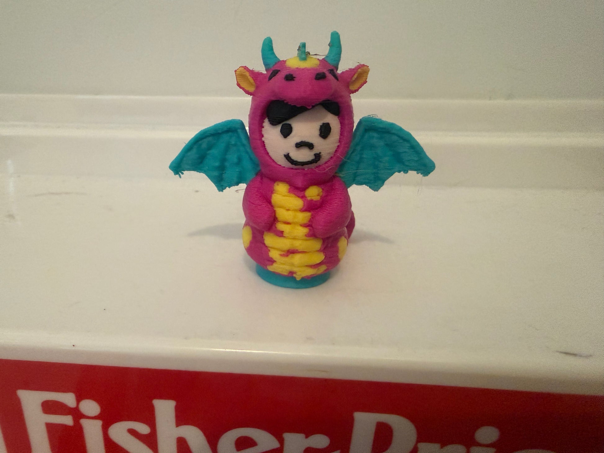 Pink dragon costume plush toy with teal wings and yellow belly, front-facing smile.