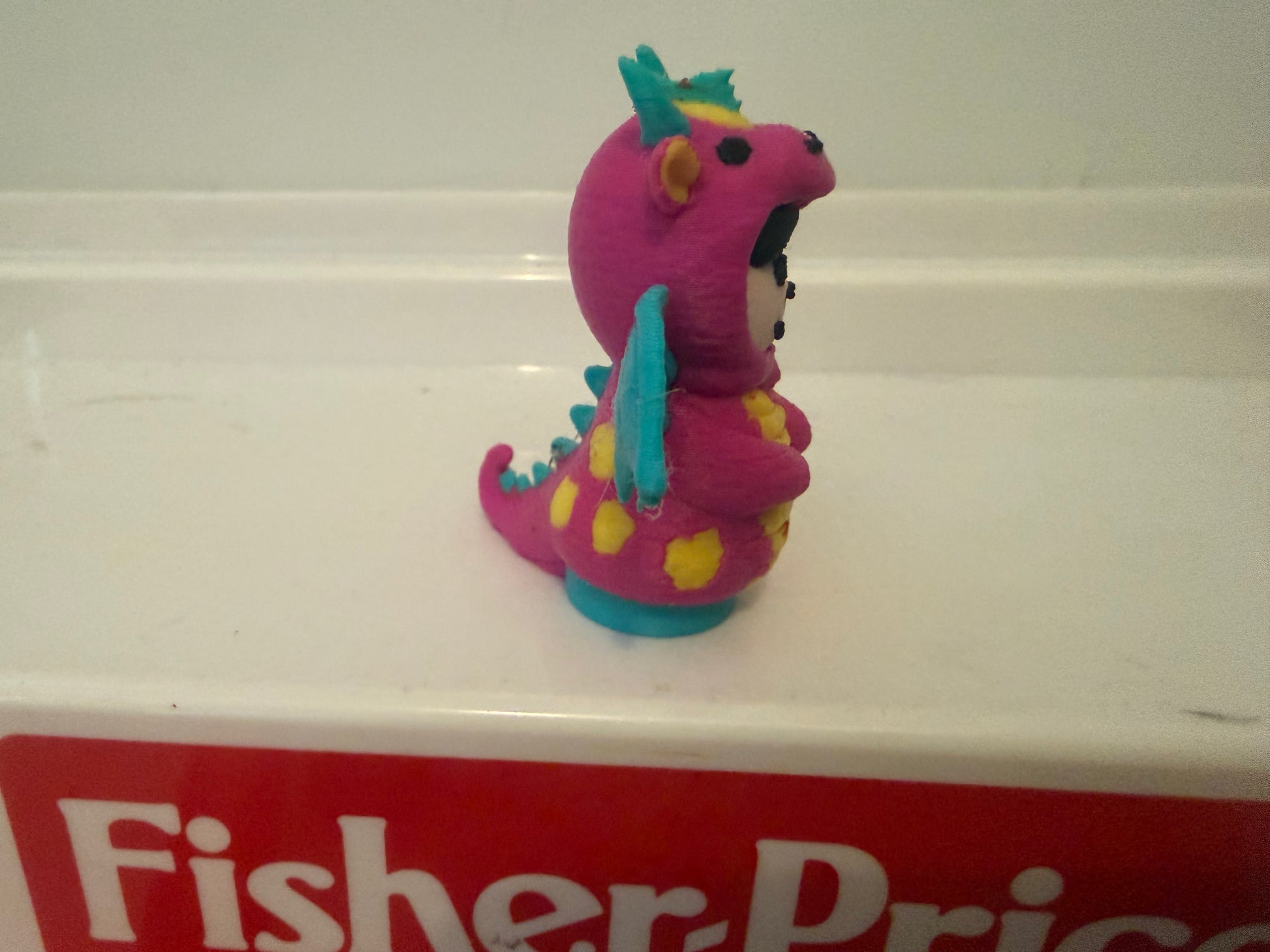 Pink dragon costume plush toy in profile, teal wings, yellow spots, smiling face.