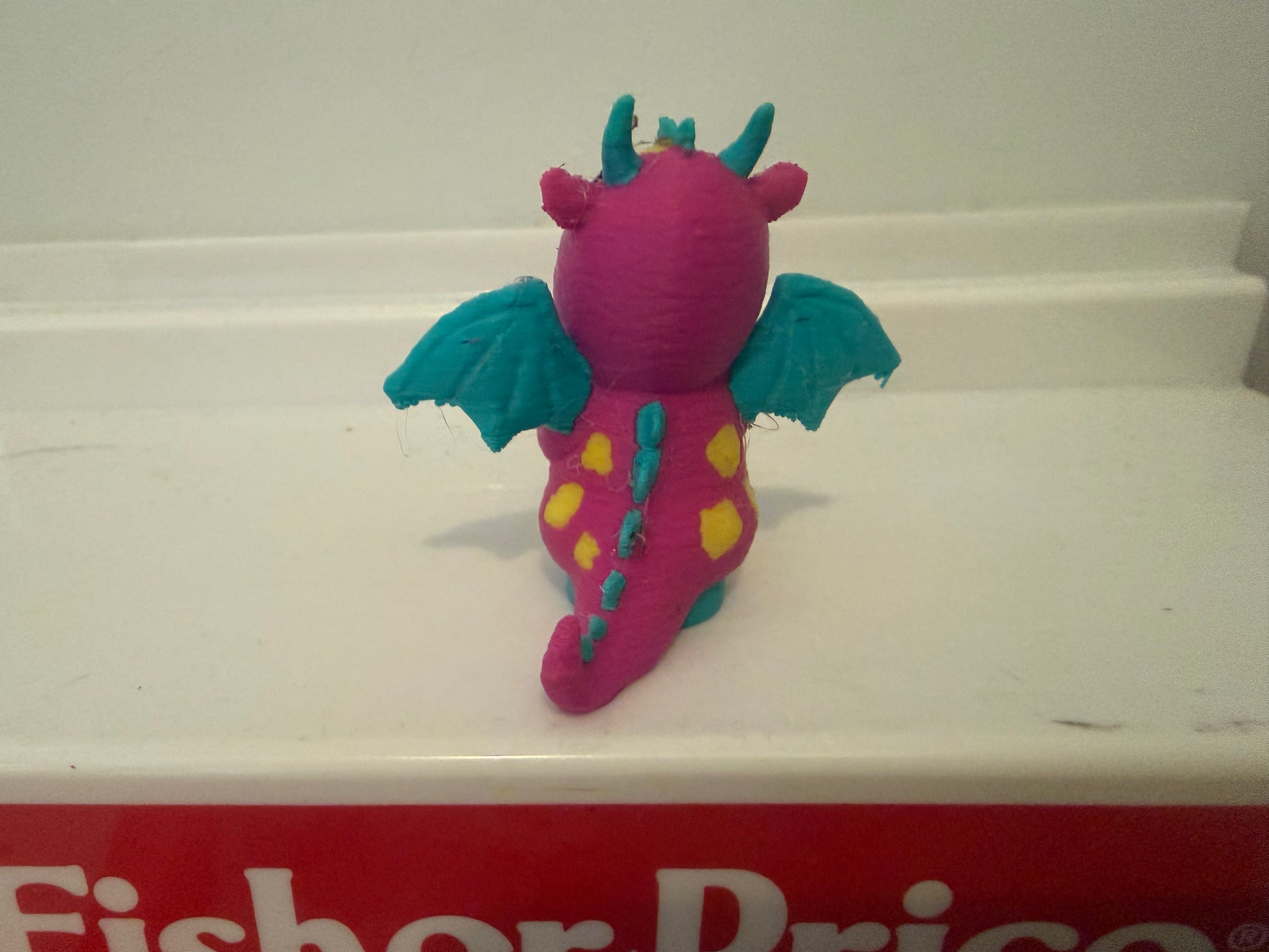 Pink dragon costume plush toy from behind with teal wings and long blue-green tail.