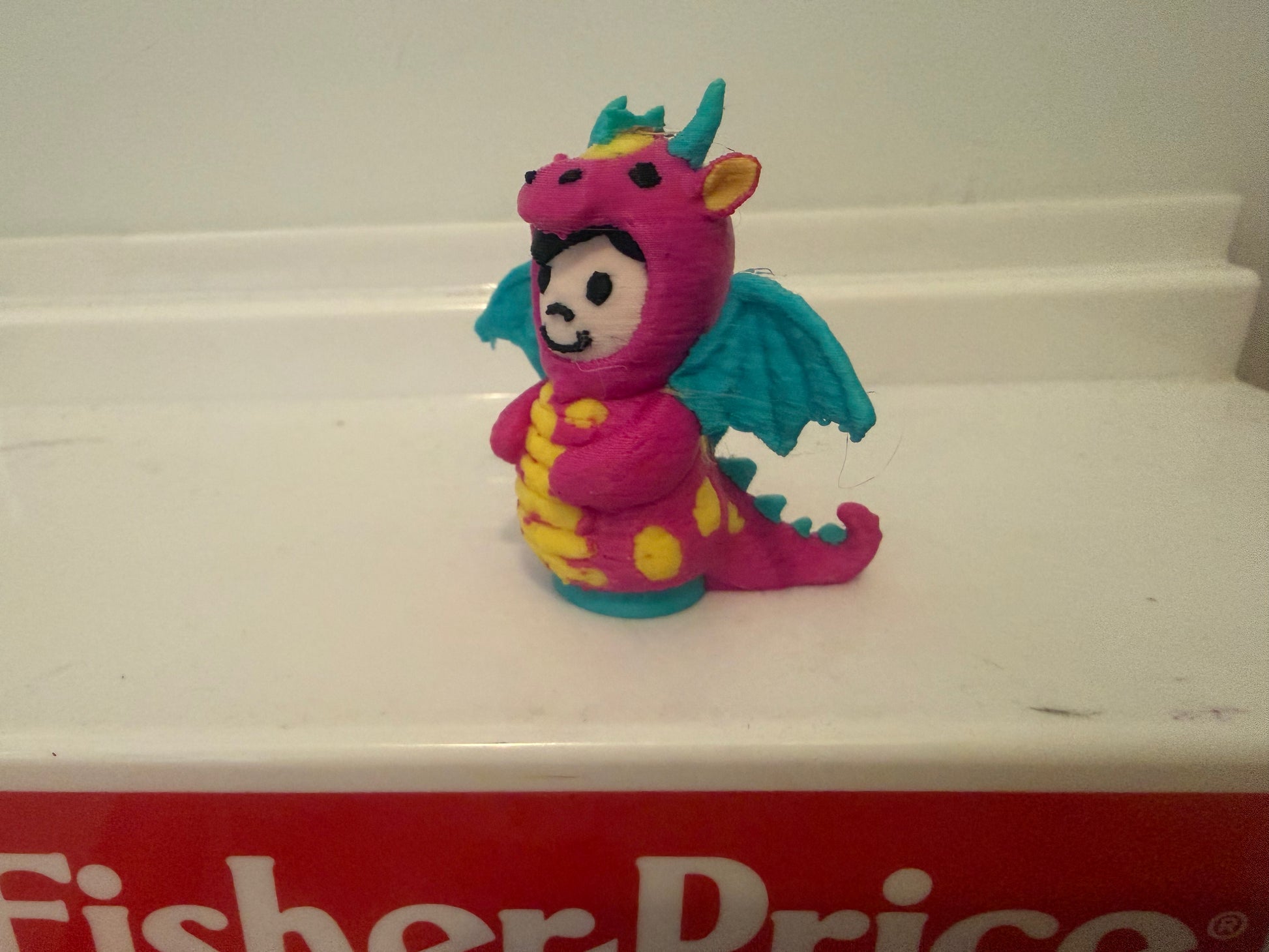 Pink dragon costume plush toy seated with teal wings and curled tail, side view.