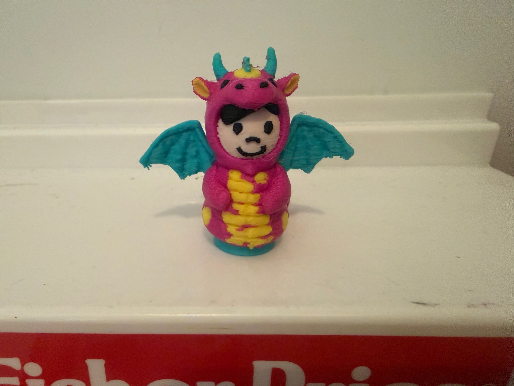 Pink dragon costume plush toy with teal wings and yellow belly, smiling face.