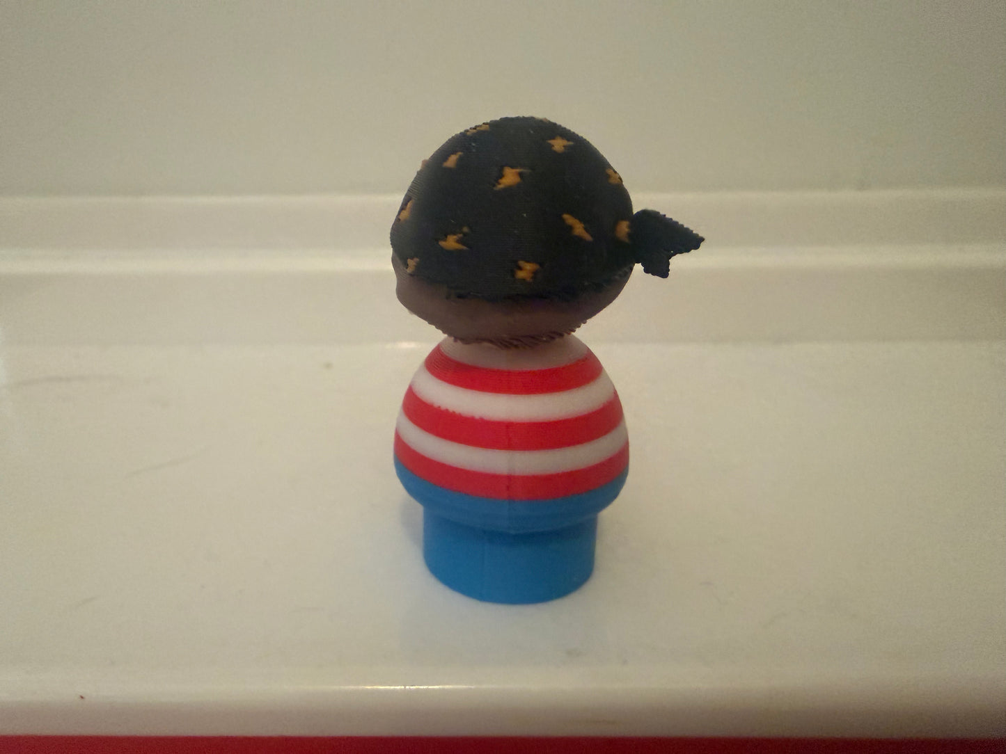 Pirate toy figure side view: black tricorn hat with gold accents, striped shirt, blue base