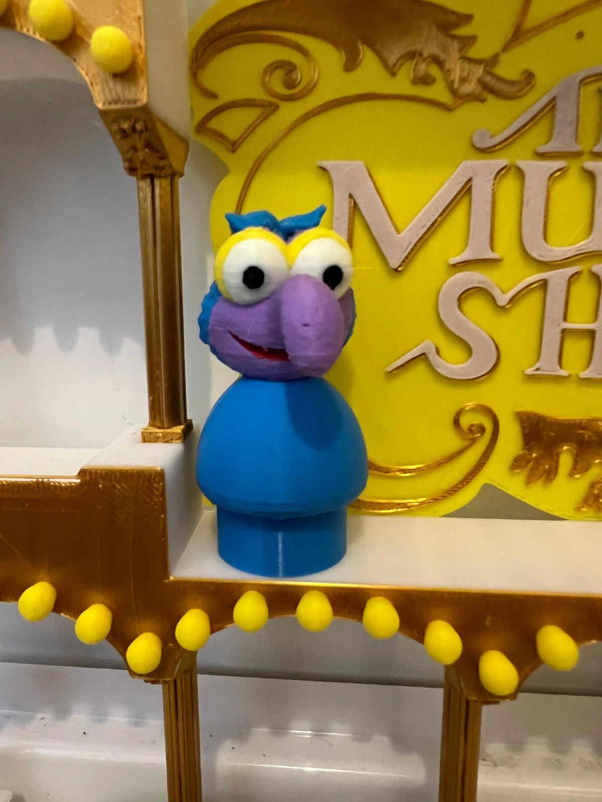 A custom 3D-printed Muppets-inspired Little People figure of Gonzo, in blue and purple, stands before a yellow sign.