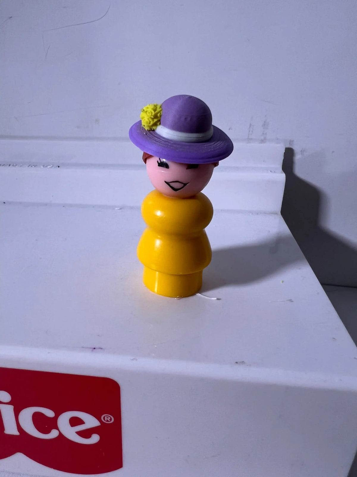 A custom Fisher-Price Little People figure wears a fancy summer hat with a purple brim and white band, adorned with a yellow flower.