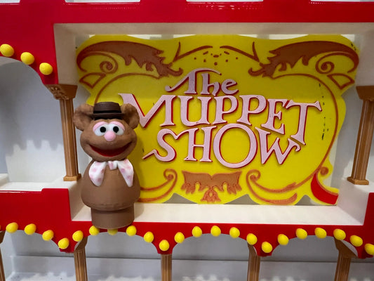 A custom Little People Fozzie Bear figure stands on a red stage in front of a yellow "The Muppet Show" sign.