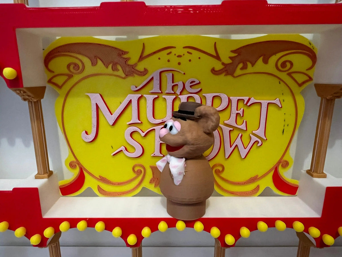 Fozzie Bear puppet figure with raised hand in front of yellow "The Muppet Show" sign