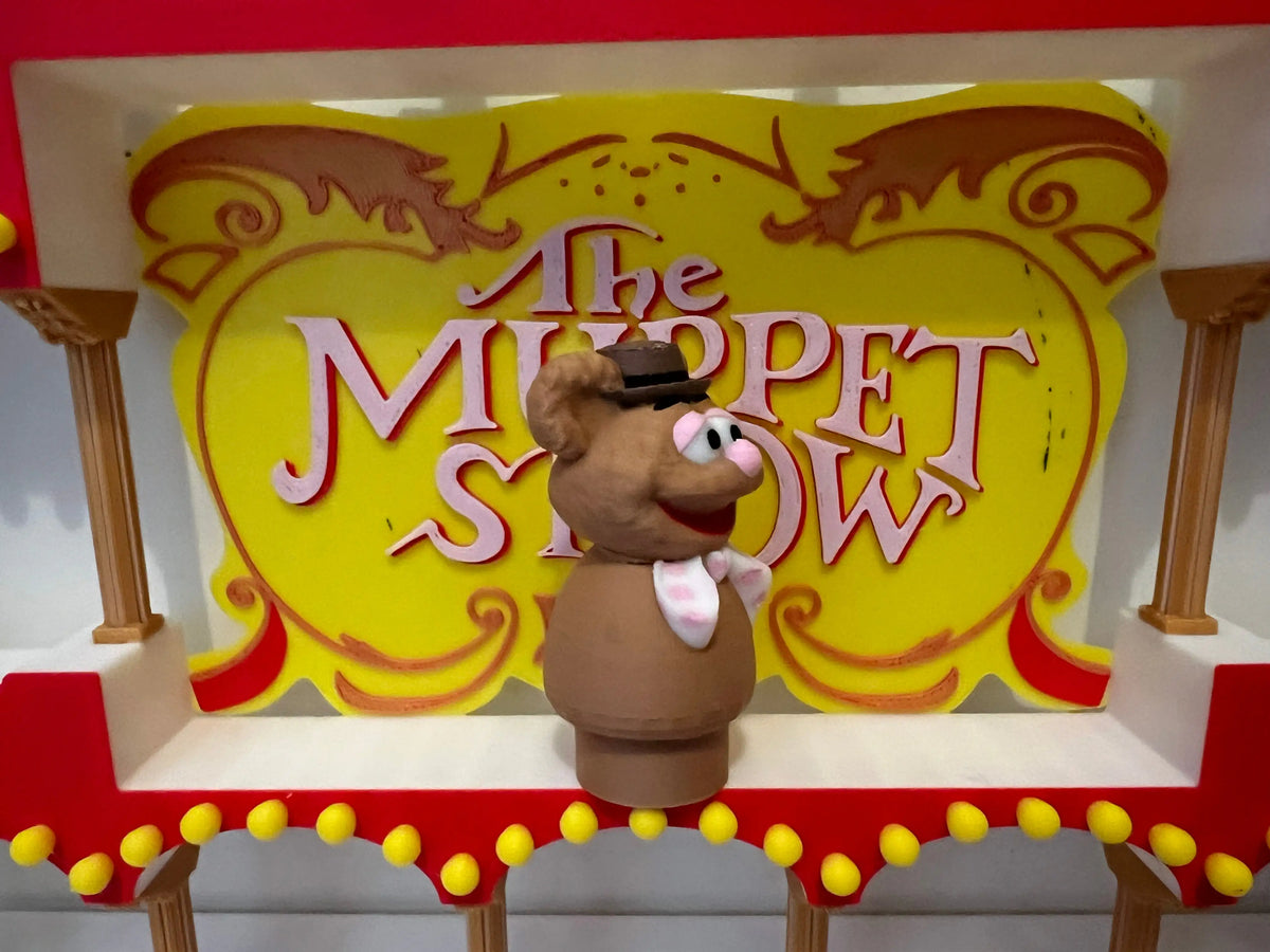 Fozzie Bear puppet figure smiling in front of yellow "The Muppet Show" sign