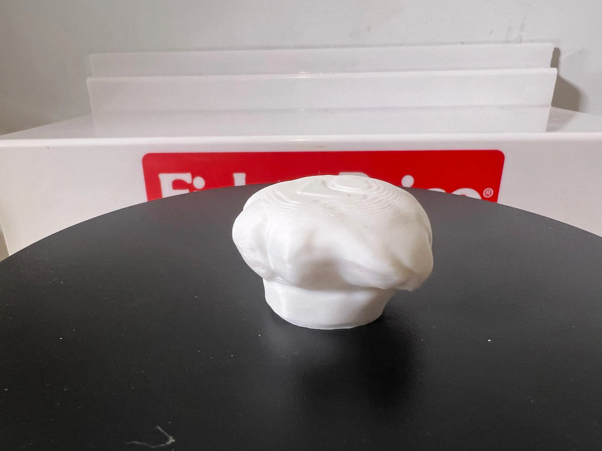Miniature white chef hat with gathered top, close-up on dark pedestal