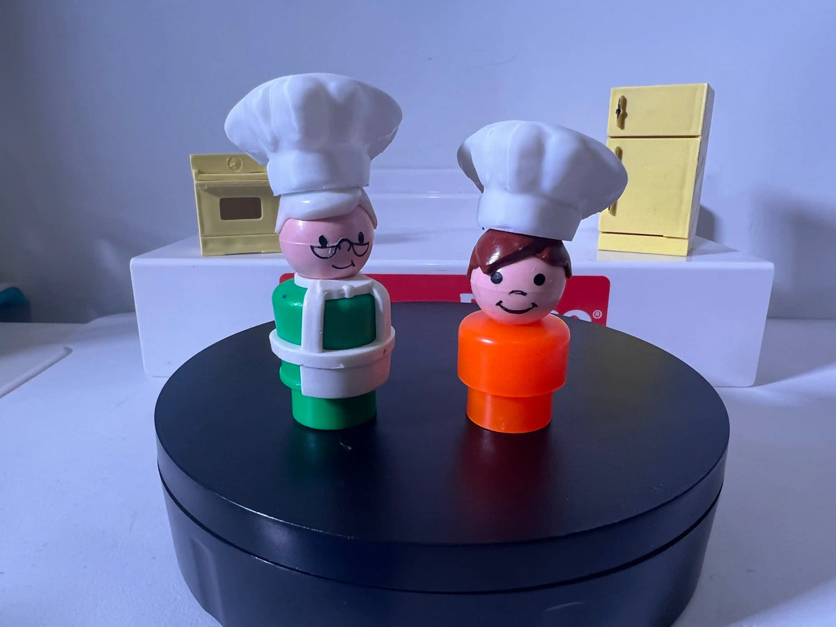 Two custom Little People scale figures wearing miniature chef hats stand before a toy oven and refrigerator.