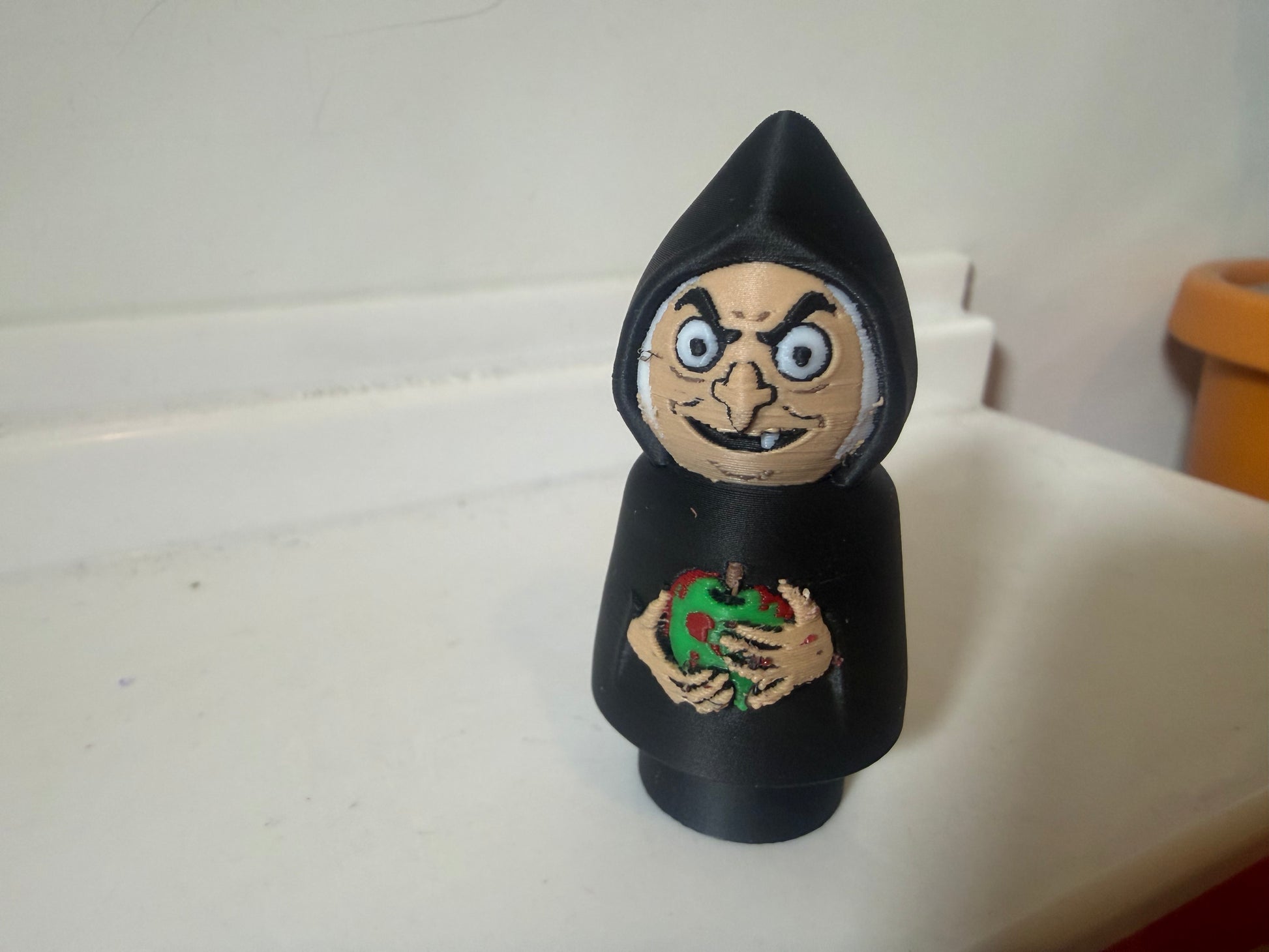 Hooded villain figure in black with wide eyes, painted face, and chest emblem