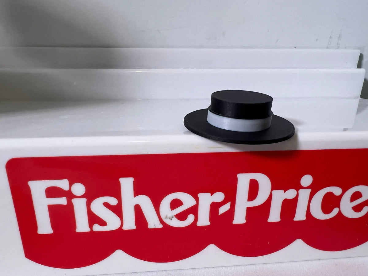 A black custom miniature designer hat with a gray band for vintage Fisher-Price Little People figures, on a Fisher-Price box.