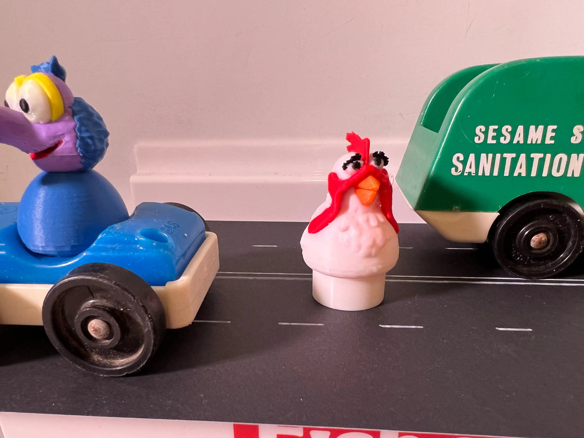 A custom 3D printed Little People Camilla Muppet figure stands on a road next to a green Sesame Street Sanitation truck.