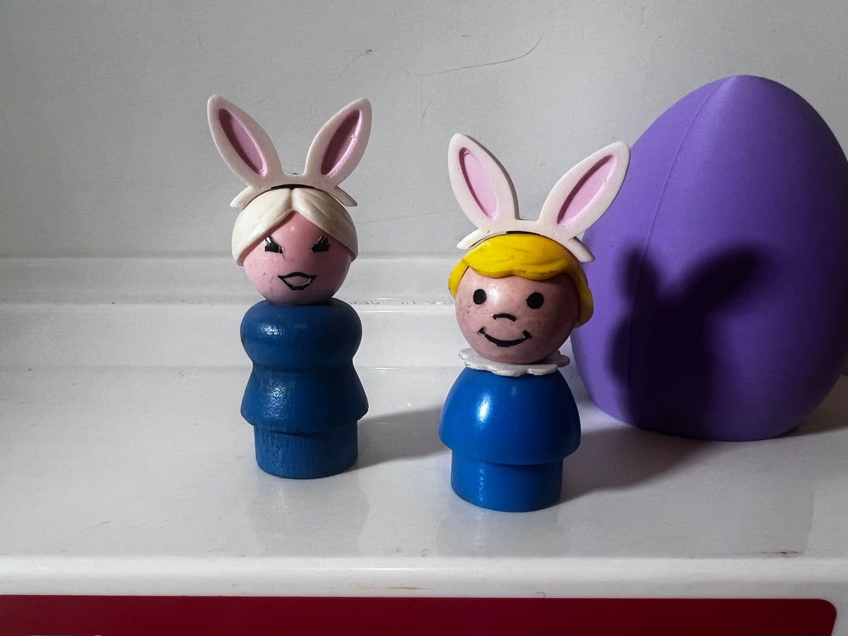 Two custom Fisher-Price Little People figures wearing bunny ear headbands stand next to a large purple Easter egg.