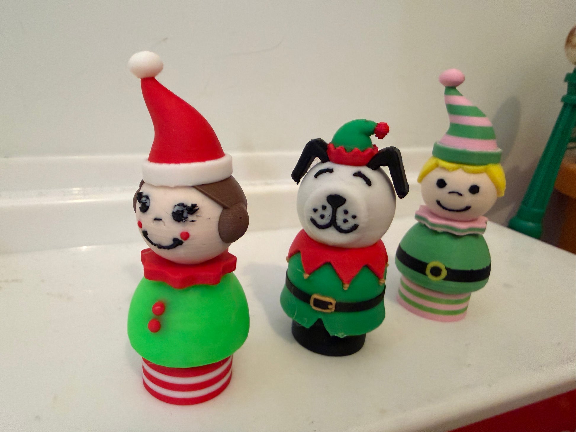 Three standing holiday figurines on a shelf: dog elf, red striped elf, green striped cat