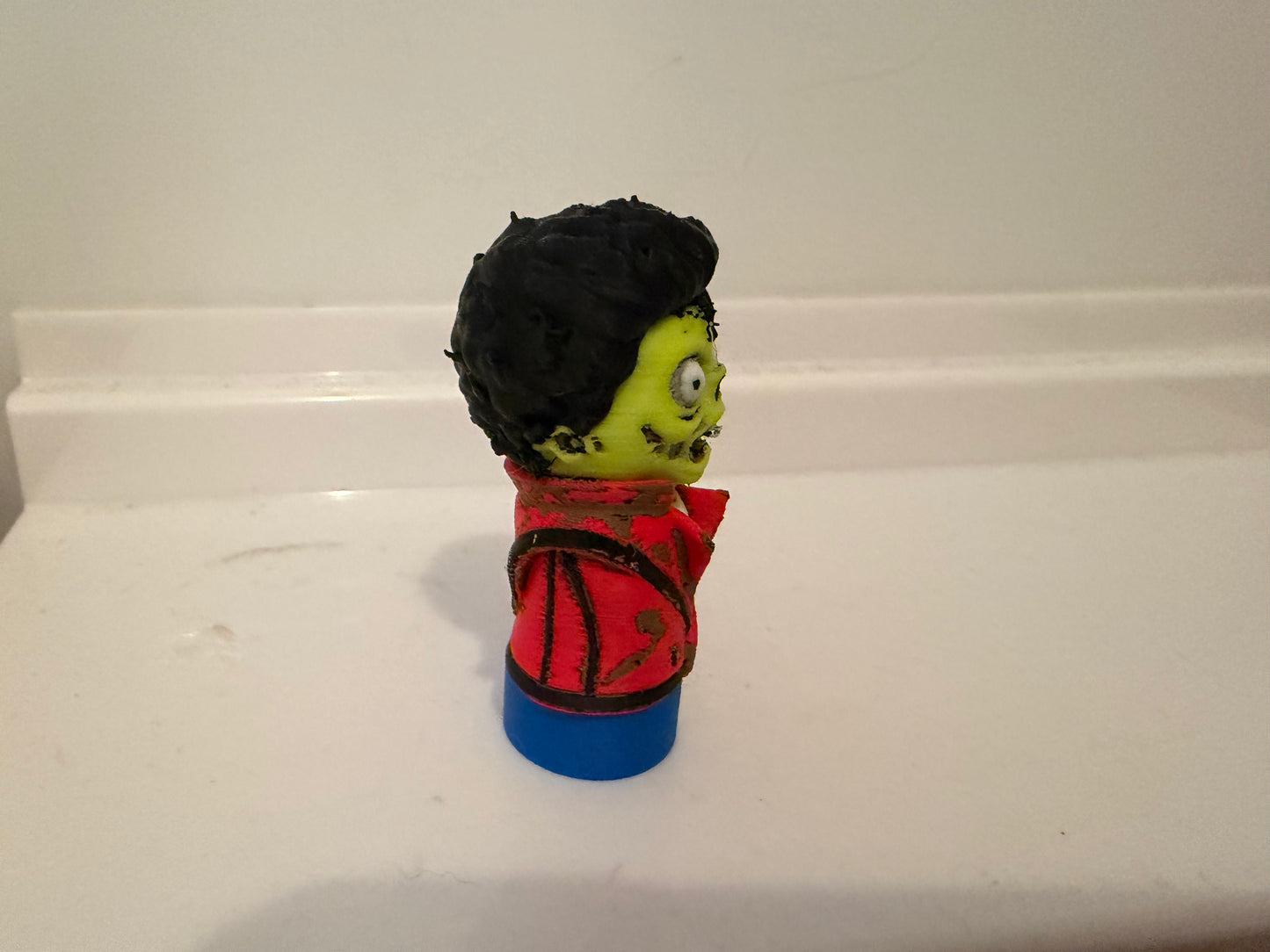 Zombie figurine side profile, green face, black hair, red jacket, blue base