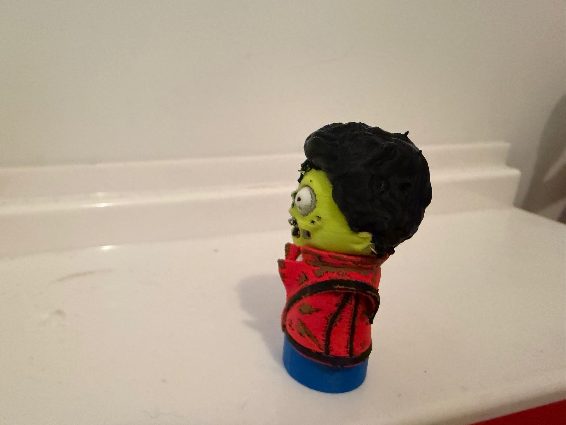 Zombie figurine angled view with green skin, black hair, red jacket, blue base
