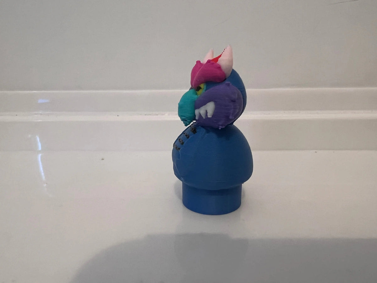 Blue monster toy with purple head, teal body, pink crest, and small arms