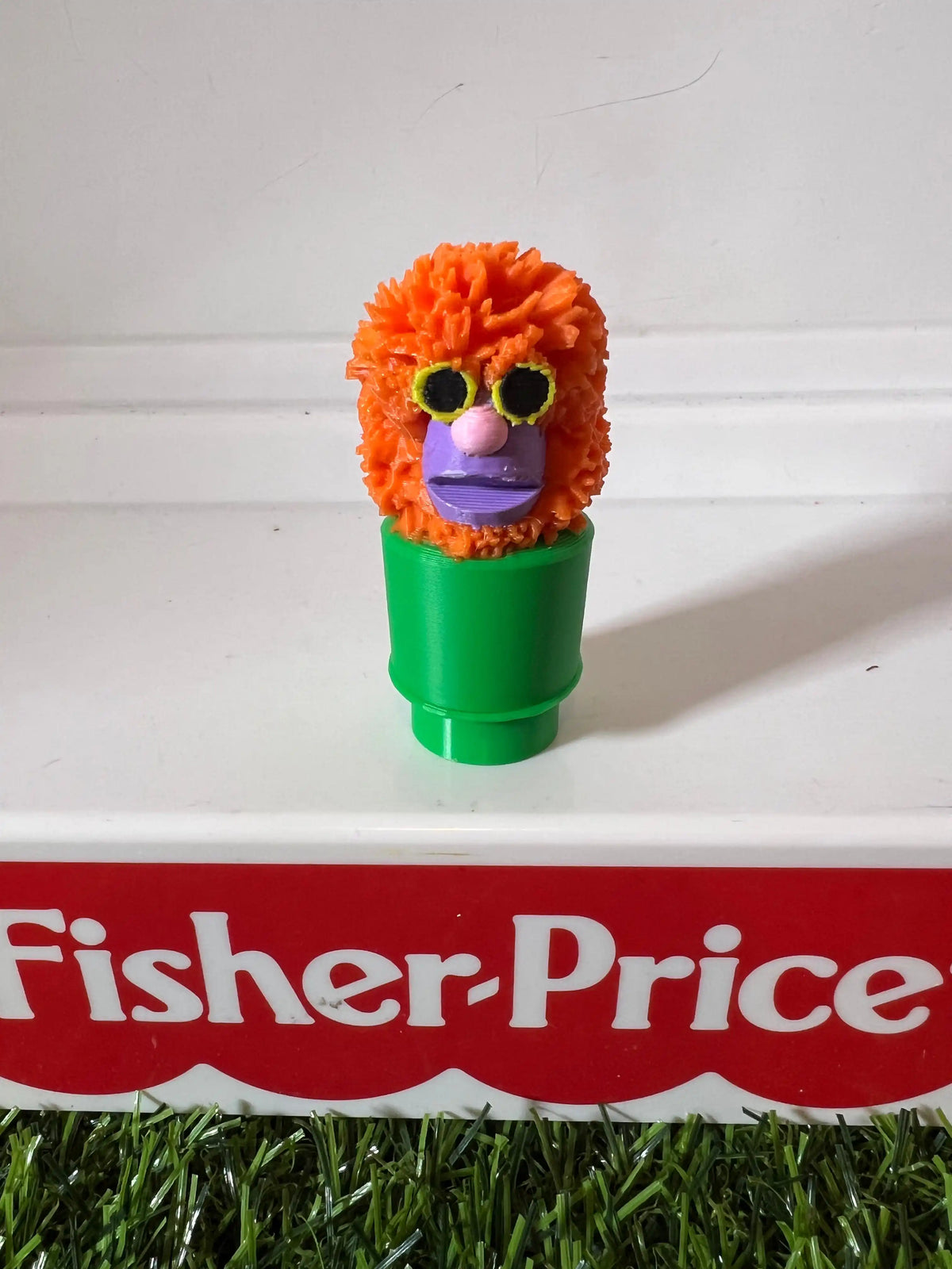 Orange fuzzy puppet with purple face and green tube base, front view.