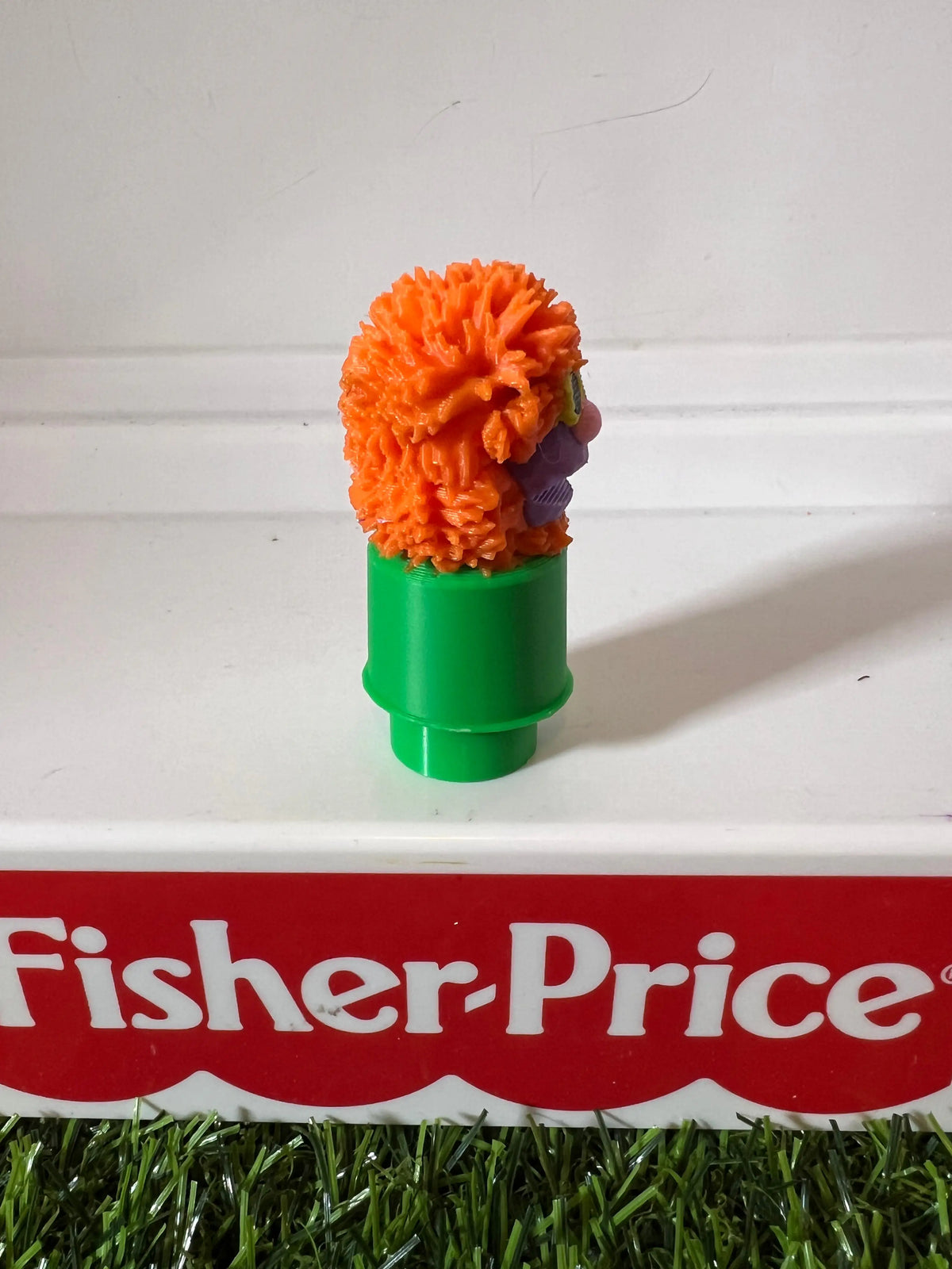 Orange fuzzy puppet on green tube base, side profile with purple face.