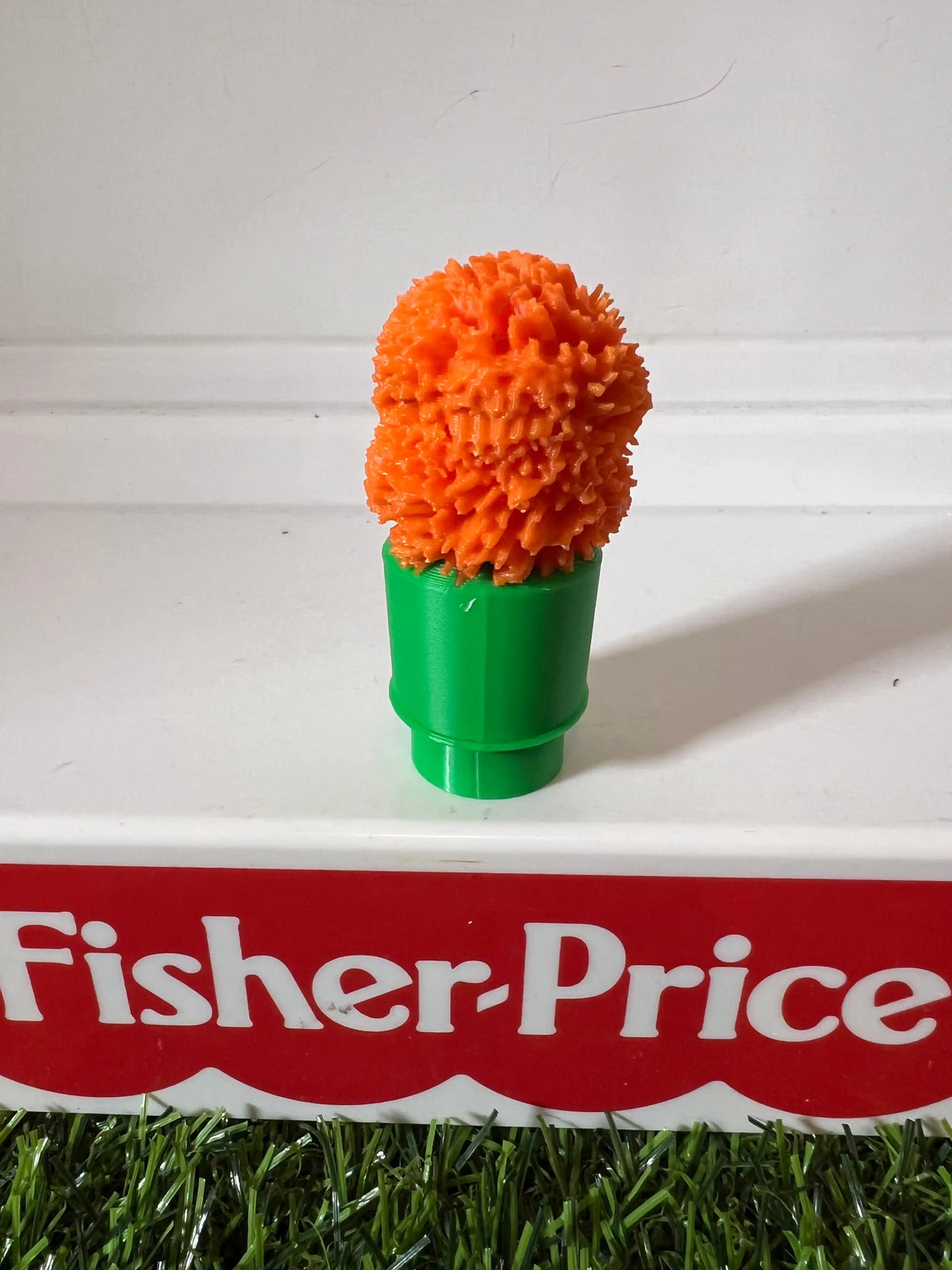 Orange fuzzy puppet head on green tube base, top-leaning front view.