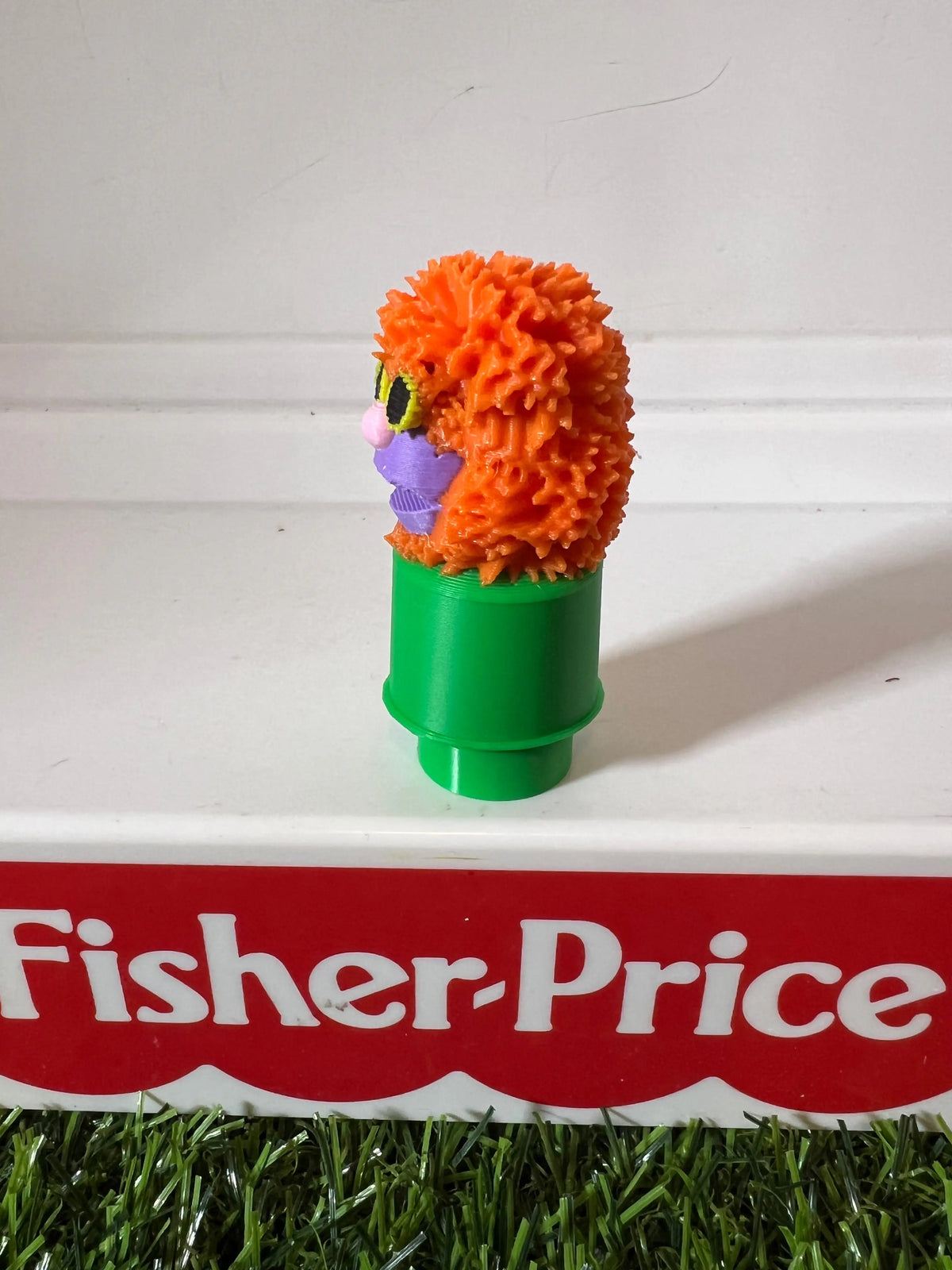 Orange fuzzy puppet with purple face and green tube base, slight side view.