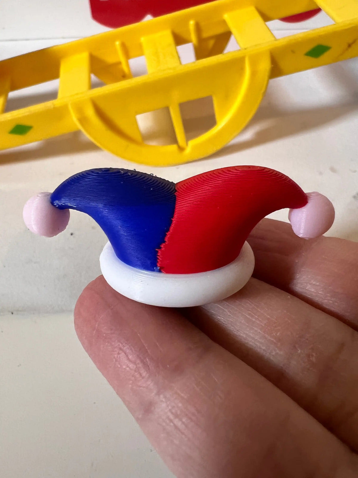 A custom 3D printed Little People miniature jester hat in red and blue with pink pompoms, held above a yellow toy.