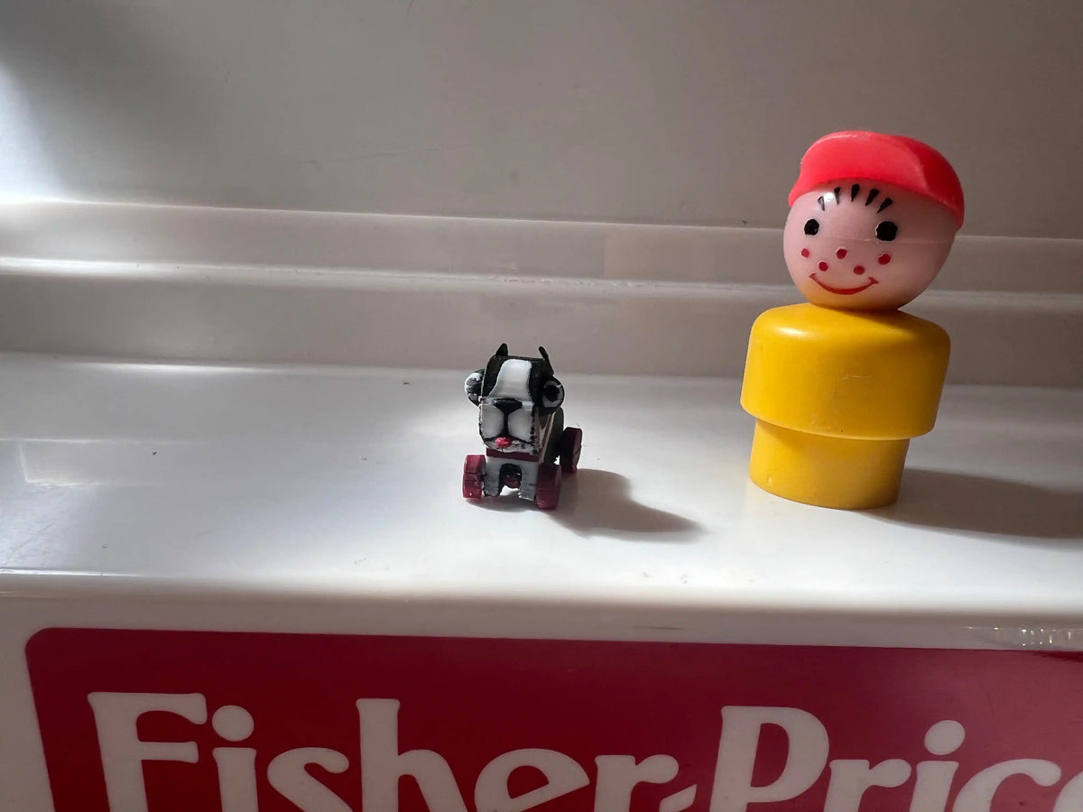 Mini child figure in red cap and yellow body next to small black dog on red Fisher-Price box
