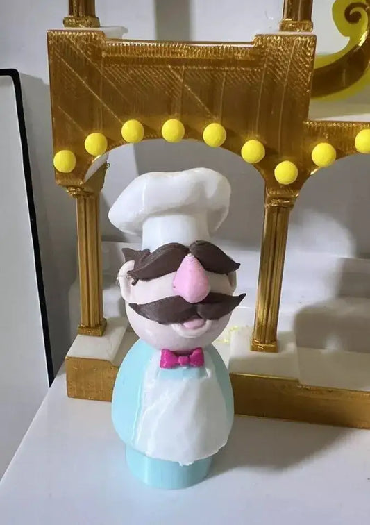 A custom 3D printed Swedish Chef Little People figure, with a pink nose and brown mustache, stands under a golden archway.