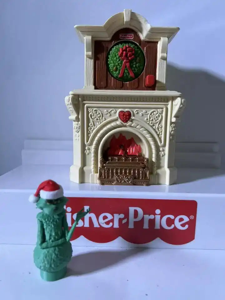 A custom 3D printed Grinch Little People figure in a Santa hat stands before a Fisher-Price fireplace.