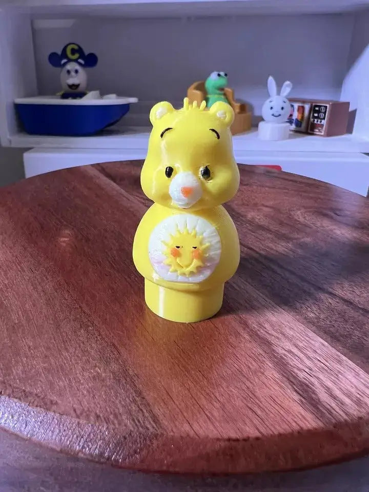 A custom yellow Fisher-Price Little People Funshine CareBear figure stands on a wooden surface with other toy figures in the background.