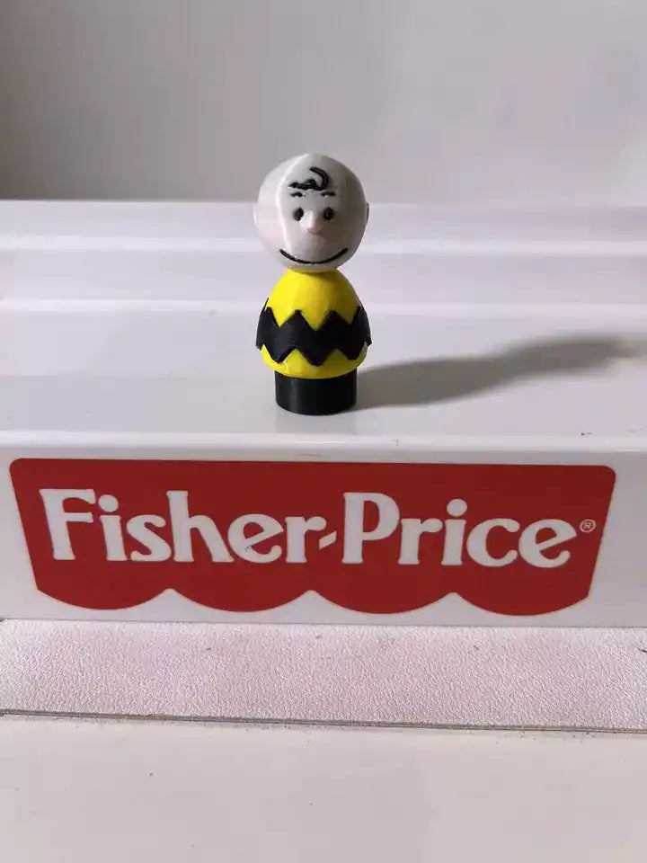 A 3D printed toy figure of Charlie Brown standing on a platform with the Fisher-Price logo.
