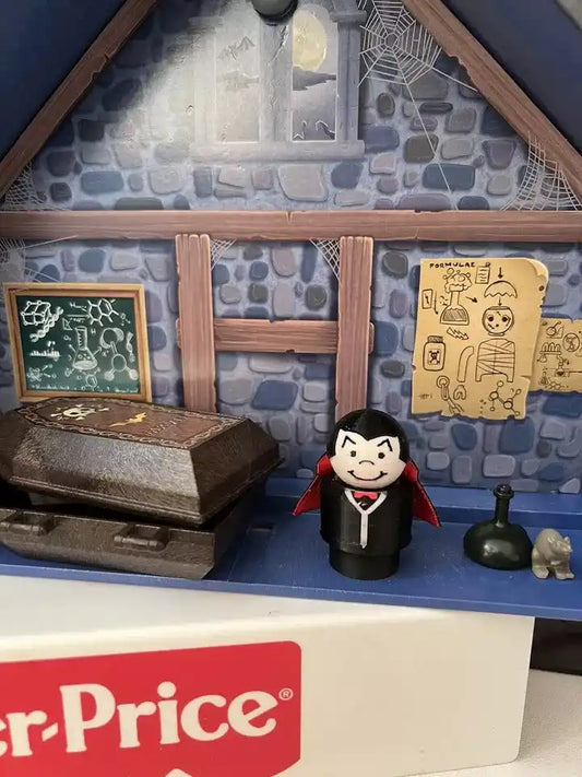 A custom Fisher-Price Little People Uncle Dracula figure with a red cape in a Halloween diorama with a coffin.
