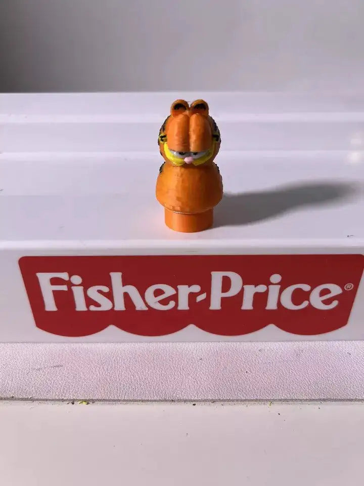 A custom 3D printed Garfield Little People figure, orange with black stripes, stands on a white surface with a red Fisher-Price logo.