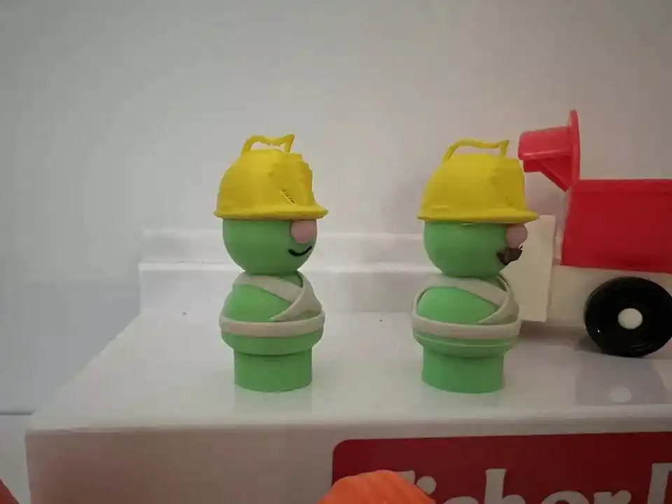 Two green Doozer figures with yellow caps posed beside a red toy cart and wheel