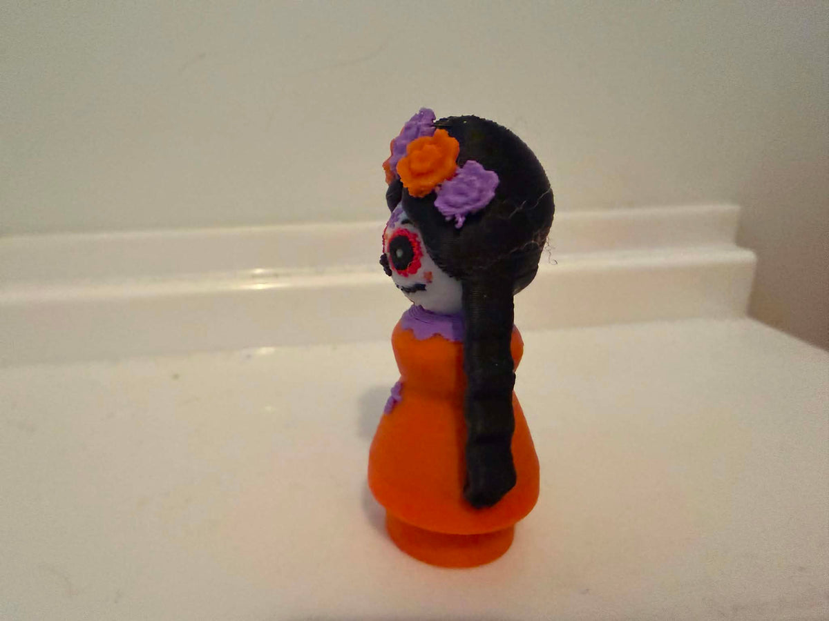 Catrina-style girl figurine angled view with orange dress and purple flower accents