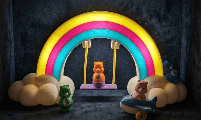 Rainbow lightbox display with glowing arch, cloud base, and hanging bear figures on swings.
