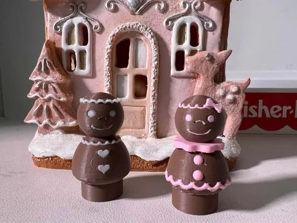 Custom 3D-printed Fisher-Price Little People-style Gingerbread Pals (boy and girl) in front of a pink gingerbread house.