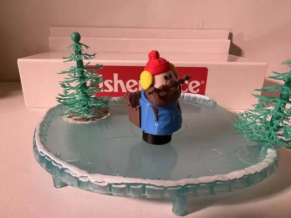 A custom 3D printed Little People-style "Yukon Cornelious" figure stands on a frozen pond with two small green trees.