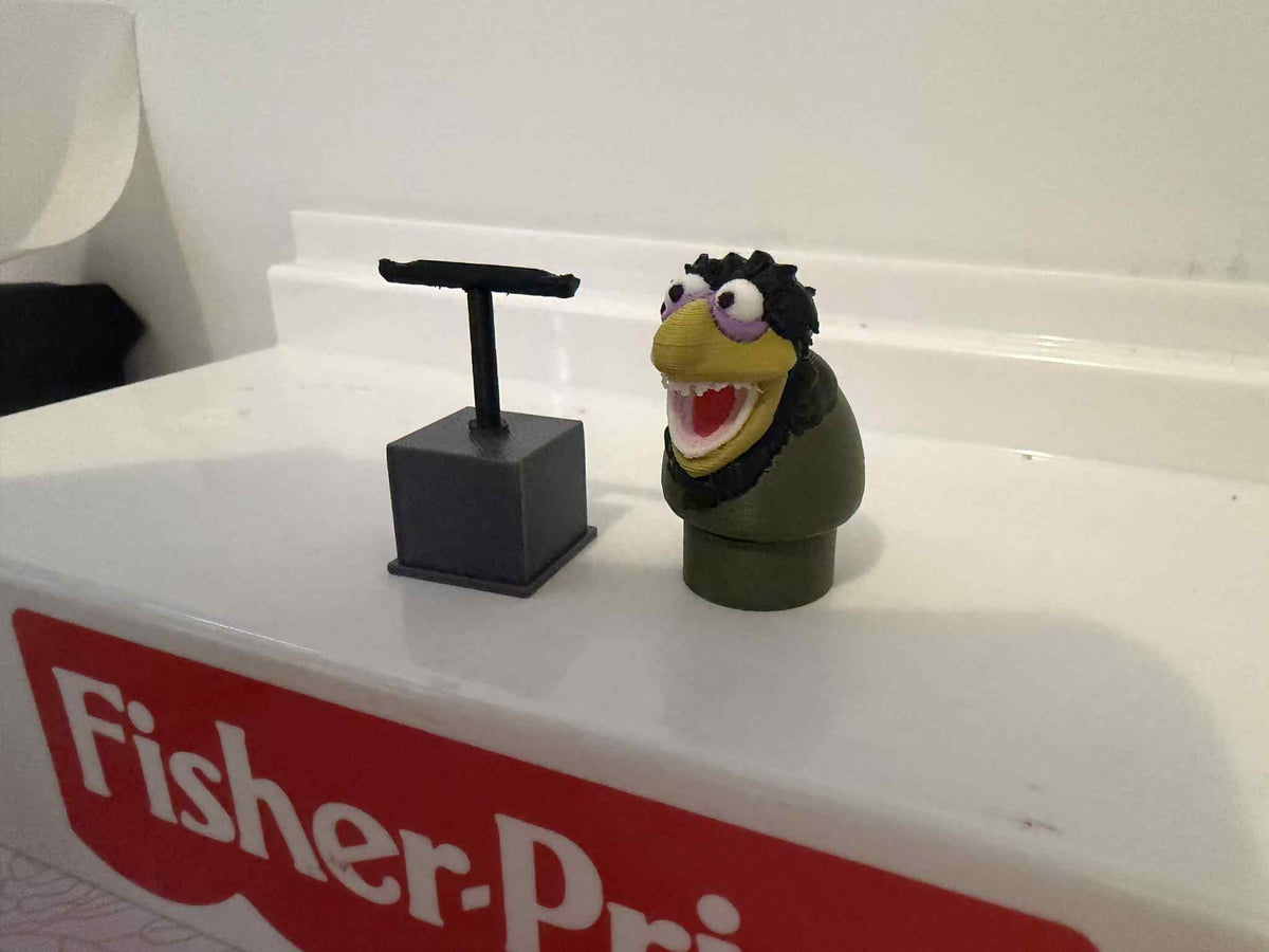 Green muppet figure with open mouth and purple glasses, next to dark stand