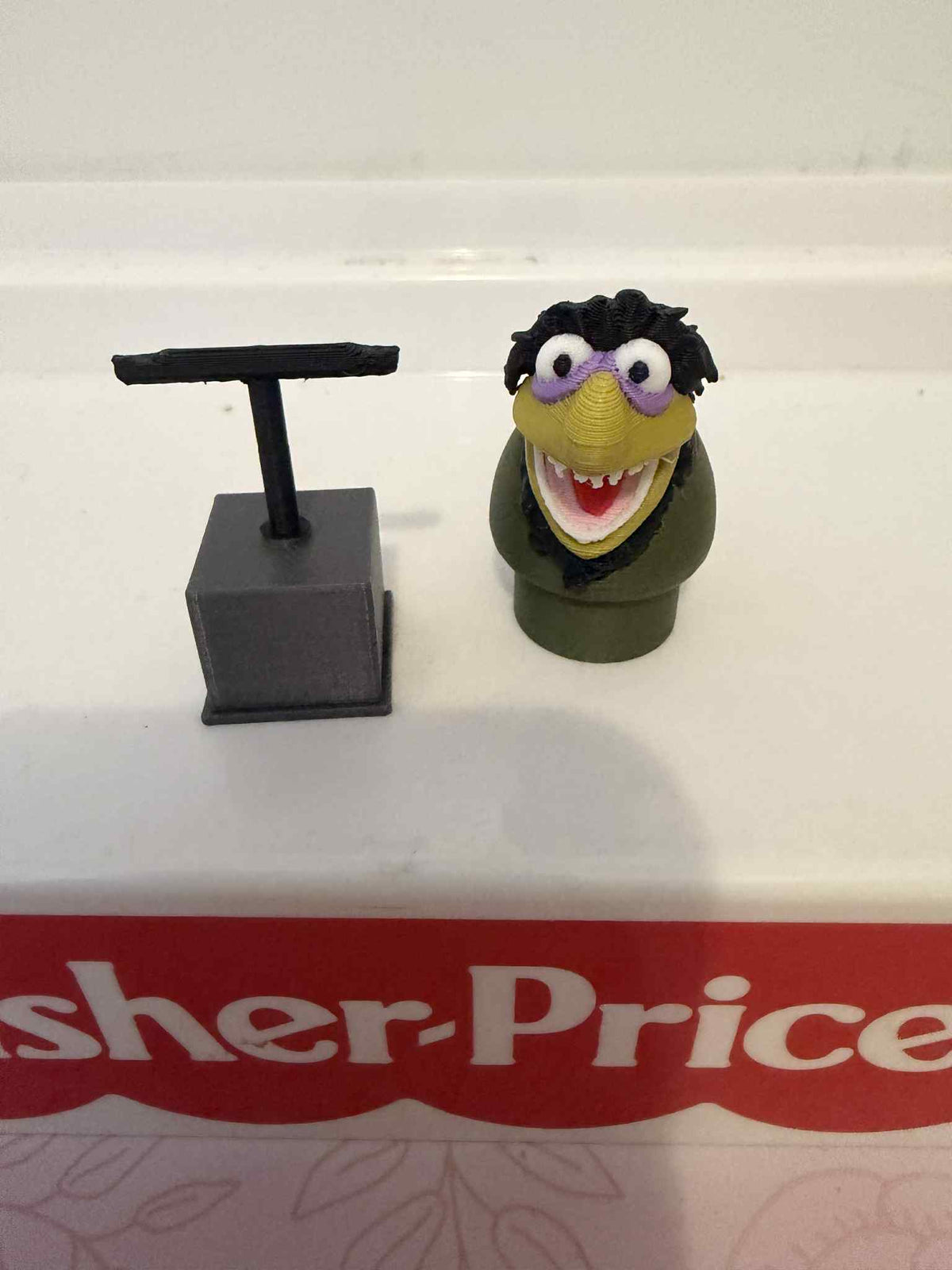 Front view of green muppet figure with purple glasses and open mouth on dark stand