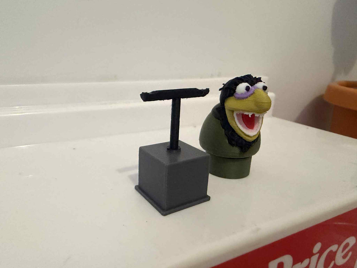 Green muppet figure with purple glasses and open mouth, on dark stand